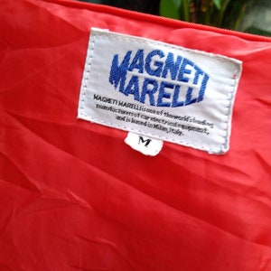 Op de afbeelding: Close-up van rode stof met een wit label. Het label toont de blauwe tekst "MAGNETI MARELLI" en de tekst "MAGNETI MARELLI is een van 's werelds toonaangevende fabrikanten van elektrische auto-uitrusting en is gevestigd in Milaan, Itali&euml;." Een maat "M" label is zichtbaar.
