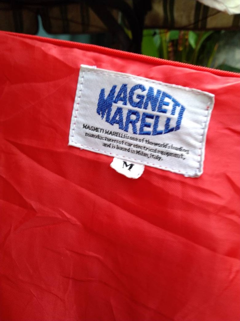 Op de afbeelding: Close-up van rode stof met een wit label. Het label toont de blauwe tekst "MAGNETI MARELLI" en de tekst "MAGNETI MARELLI is een van 's werelds toonaangevende fabrikanten van elektrische auto-uitrusting en is gevestigd in Milaan, Itali&euml;." Een maat "M" label is zichtbaar.