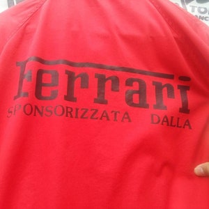 K&ouml;nnte beinhalten: Rote Jacke mit einem schwarzen Ferrari-Logo und dem Text "SPONSORIZZATA DALLA" auf der R&uuml;ckseite.