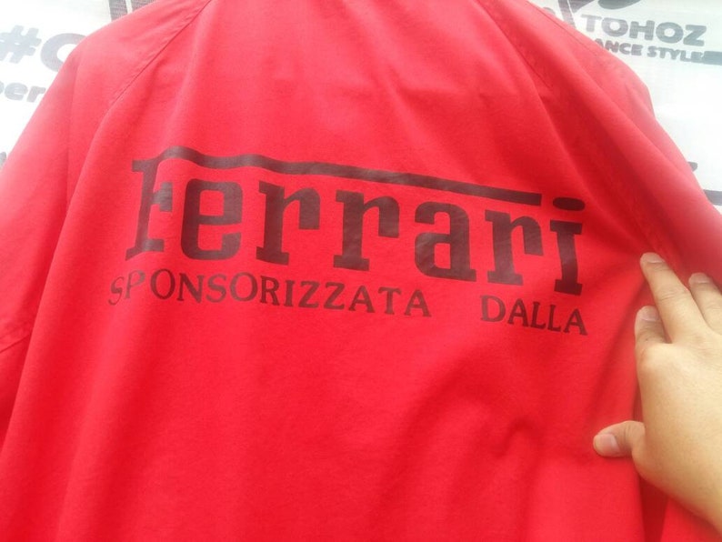 K&ouml;nnte beinhalten: Rote Jacke mit einem schwarzen Ferrari-Logo und dem Text "SPONSORIZZATA DALLA" auf der R&uuml;ckseite.