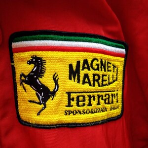 Op de afbeelding: Rode jas met een gele patch met het Ferrari-logo, een zwart steigerend paard en de tekst "MAGNETI MARELLI Ferrari". De patch heeft groene, witte en rode strepen aan de bovenkant.