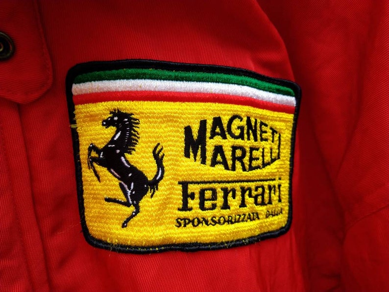 Op de afbeelding: Rode jas met een gele patch met het Ferrari-logo, een zwart steigerend paard en de tekst "MAGNETI MARELLI Ferrari". De patch heeft groene, witte en rode strepen aan de bovenkant.