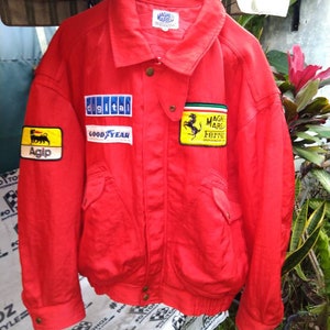Op de afbeelding: Een felrode jas met meerdere patches, waaronder "digital", "GOOD YEAR" en "Agip". De jas heeft een kraag, zakken en het "MAGNET MARELLI" logo. Een gele patch met een zwart paard is ook zichtbaar.
