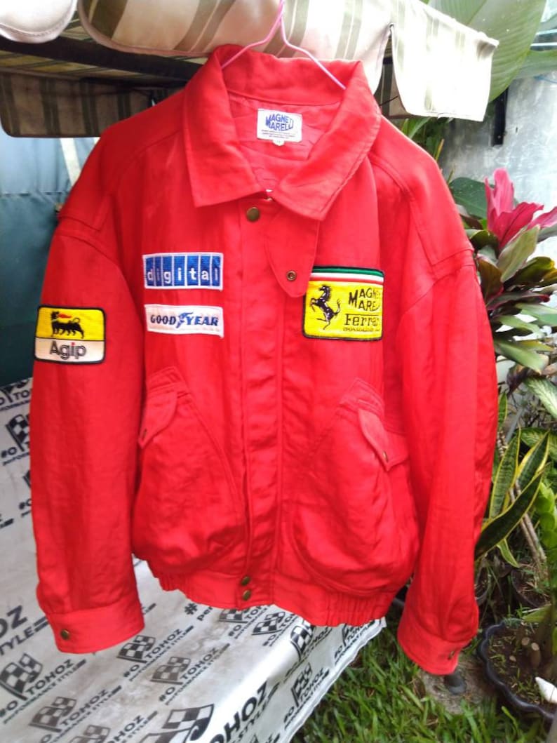 Op de afbeelding: Een felrode jas met meerdere patches, waaronder "digital", "GOOD YEAR" en "Agip". De jas heeft een kraag, zakken en het "MAGNET MARELLI" logo. Een gele patch met een zwart paard is ook zichtbaar.