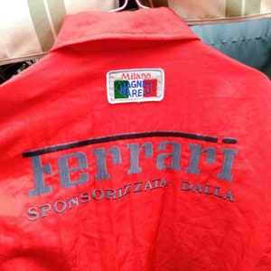 Op de afbeelding: Rode jas met het woord "Ferrari" in grijs op de rug geborduurd. De jas heeft ook de tekst "SPONSORIZZATA DALLA" en een patch met "MILANO MAGNETI MARELLI".