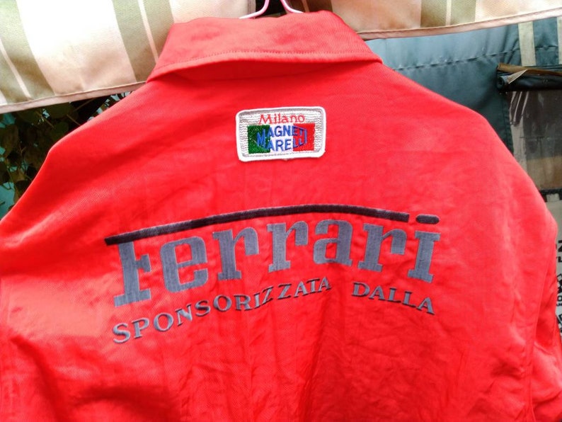 Op de afbeelding: Rode jas met het woord "Ferrari" in grijs op de rug geborduurd. De jas heeft ook de tekst "SPONSORIZZATA DALLA" en een patch met "MILANO MAGNETI MARELLI".