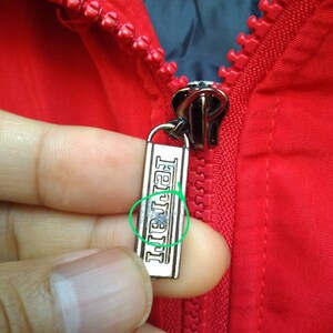 Puede incluir: Una cremallera roja con un tirador de cremallera plateado. El tirador de la cremallera tiene la palabra "Ferrari" grabada.