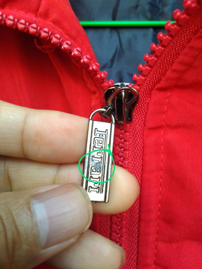 Puede incluir: Una cremallera roja con un tirador de cremallera plateado. El tirador de la cremallera tiene la palabra "Ferrari" grabada.