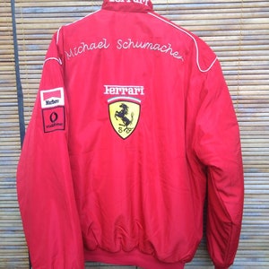 Puede incluir: Chaqueta roja Ferrari con ribetes blancos y el nombre "Michael Schumacher" bordado en la espalda. La chaqueta tiene un logotipo de Ferrari y los patrocinadores Marlboro y Vodafone.