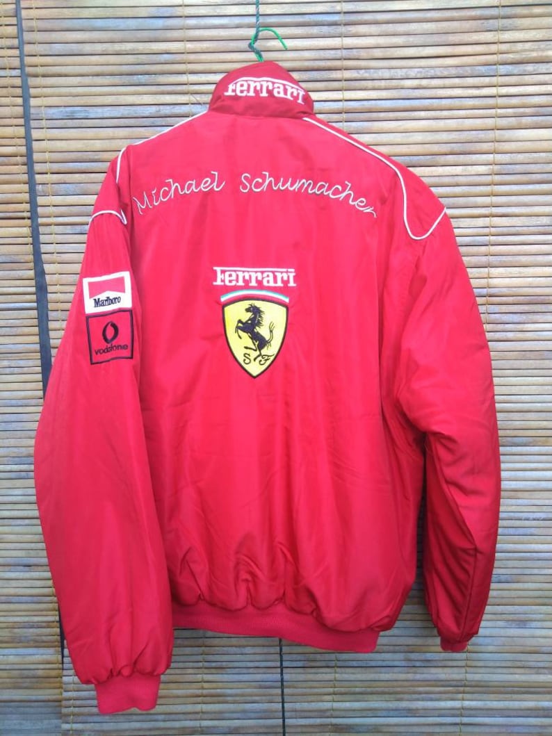 Puede incluir: Chaqueta roja Ferrari con ribetes blancos y el nombre "Michael Schumacher" bordado en la espalda. La chaqueta tiene un logotipo de Ferrari y los patrocinadores Marlboro y Vodafone.
