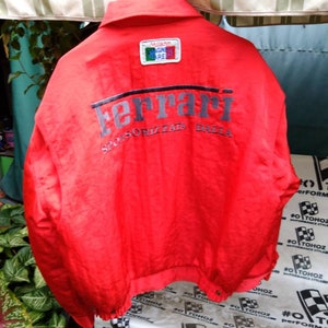 Op de afbeelding: Felrood Ferrari-jack met het grijze Ferrari-logo op de rug. De jas heeft een kleine patch met de kleuren van de Italiaanse vlag en de woorden "Milano" en "Magna Pare". De tekst "SPONSORIZZATA DALLA" staat onder het Ferrari-logo.