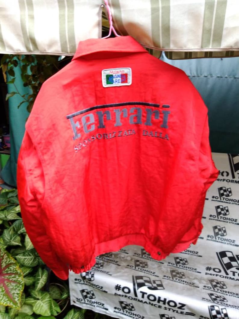 Op de afbeelding: Felrood Ferrari-jack met het grijze Ferrari-logo op de rug. De jas heeft een kleine patch met de kleuren van de Italiaanse vlag en de woorden "Milano" en "Magna Pare". De tekst "SPONSORIZZATA DALLA" staat onder het Ferrari-logo.