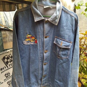 May include: A blue denim jacket with a cartoon character embroidered on the left chest. The character is a dog wearing a racing suit and holding a checkered flag. The jacket has a button-up front and two pockets.