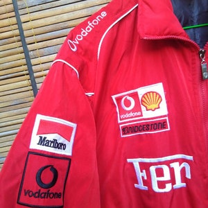Puede incluir: Chaqueta roja con logos blancos y negros. Los logos incluyen "Marlboro", "Vodafone", "Bridgestone" y "Ferrari".