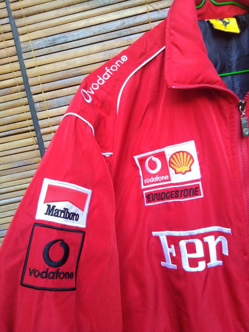 Puede incluir: Chaqueta roja con logos blancos y negros. Los logos incluyen "Marlboro", "Vodafone", "Bridgestone" y "Ferrari".