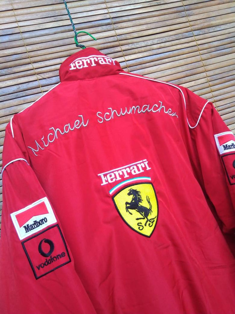 Puede incluir: Chaqueta de carreras Ferrari roja con ribetes blancos, con el logotipo de Ferrari con un caballo rampante, y los nombres "Michael Schumacher" y "Ferrari" bordados en la espalda. La chaqueta tambi&eacute;n tiene un logotipo de Marlboro y Vodafone en la manga izquierda.