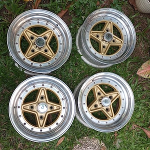 Op de afbeelding: Vier gouden en zilveren lichtmetalen velgen met een vijfpuntige stervorm. De velgen zijn geplaatst tegen een achtergrond van groen gras.