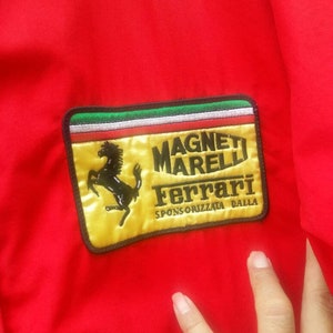 K&ouml;nnte beinhalten: Eine rote Jacke mit einem gelben Patch, auf dem "Magneti Marelli Ferrari Sponsorizzata Dalla" steht. Der Patch zeigt ein schwarzes Pferdelogo.