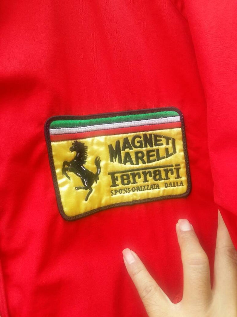 K&ouml;nnte beinhalten: Eine rote Jacke mit einem gelben Patch, auf dem "Magneti Marelli Ferrari Sponsorizzata Dalla" steht. Der Patch zeigt ein schwarzes Pferdelogo.