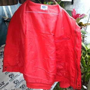 Op de afbeelding: Felrode jas met ritssluiting, elastische manchetten en een klein zakje. De jas hangt aan een roze hanger. De merknaam "MAGNE MARELLI" is zichtbaar op een label aan de binnenkant.