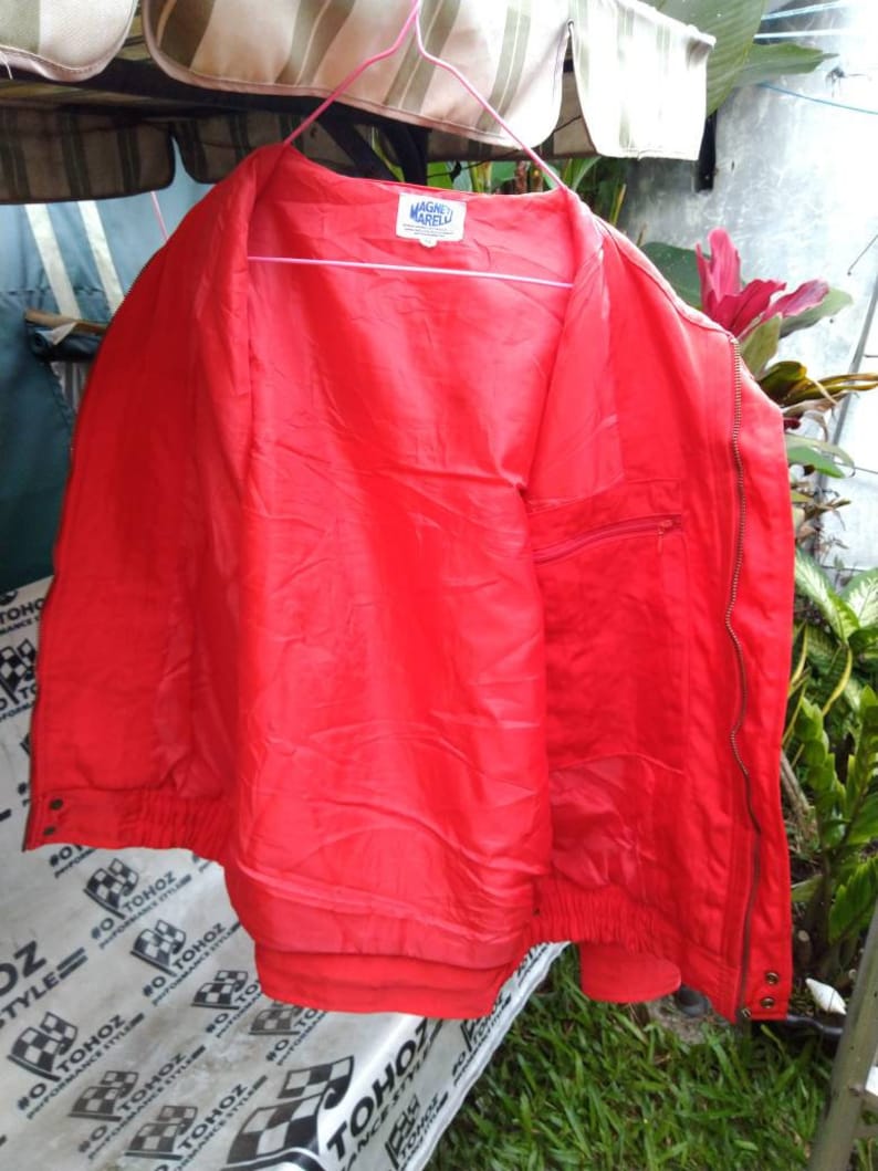 Op de afbeelding: Felrode jas met ritssluiting, elastische manchetten en een klein zakje. De jas hangt aan een roze hanger. De merknaam "MAGNE MARELLI" is zichtbaar op een label aan de binnenkant.