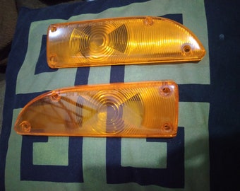 May include: Two amber-coloured plastic car turn signal lights. The lights are rectangular with rounded corners and have a circular lens with a ribbed pattern.