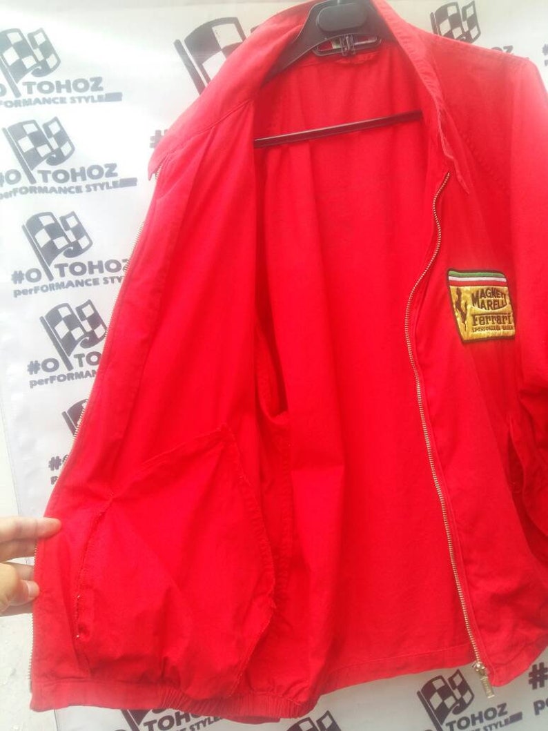 K&ouml;nnte beinhalten: Rote Windbreaker-Jacke mit Rei&szlig;verschluss und einem kleinen gestickten Aufn&auml;her auf der linken Brust, auf dem "Magni Marelli Ferrari" steht.