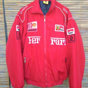 Puede incluir: Chaqueta roja Ferrari con detalles blancos y logotipos de patrocinadores, incluyendo Vodafone, Marlboro y Bridgestone. La chaqueta tiene cierre de cremallera y dos bolsillos.