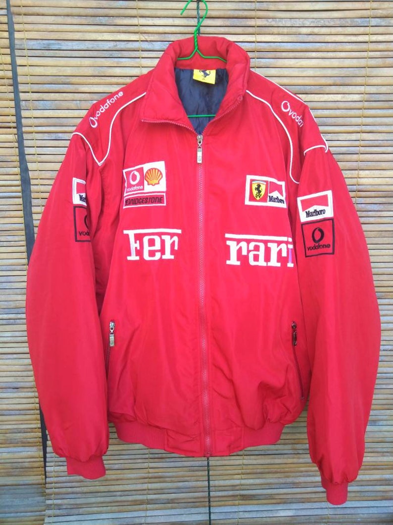 Puede incluir: Chaqueta roja Ferrari con detalles blancos y logotipos de patrocinadores, incluyendo Vodafone, Marlboro y Bridgestone. La chaqueta tiene cierre de cremallera y dos bolsillos.