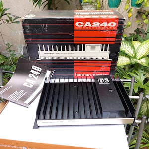 May include: A black Harman Kardon CA240 high fidelity car amplifier with a silver faceplate and a black box with the text "CA240 High Fidelity Car Amplifier" on it.