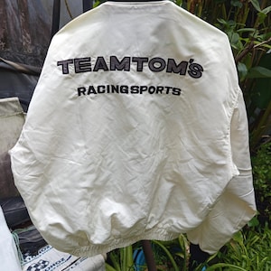 May include: Cream-colored racing jacket with black ribbed collar and cuffs. The back of the jacket features the embroidered text "TEAM TOM'S RACING SPORTS" in black. The jacket has a classic bomber style.