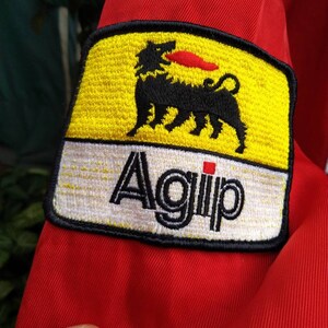 Op de afbeelding: Close-up van rode stof met een geborduurde patch. De patch is geel met een zwart, hondachtig wezen en een rode vlam. Het woord "Agip" staat in zwarte letters op een witte achtergrond.