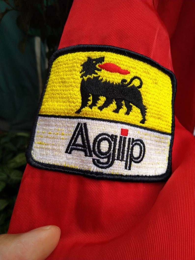 Op de afbeelding: Close-up van rode stof met een geborduurde patch. De patch is geel met een zwart, hondachtig wezen en een rode vlam. Het woord "Agip" staat in zwarte letters op een witte achtergrond.