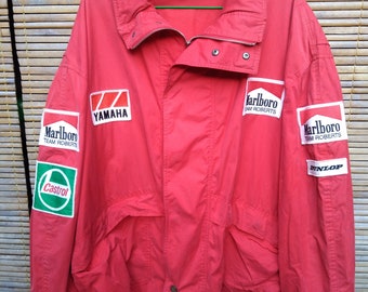 May include: A red jacket with multiple sponsor logos, including "Marlboro Team Roberts", "Yamaha", "Castrol", and "Dunlop". The jacket has a collared neckline, a zipper, and button closures. The jacket is hanging on a green hanger.