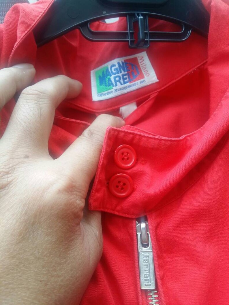 K&ouml;nnte beinhalten: Rote Jacke mit Rei&szlig;verschluss und zwei Kn&ouml;pfen. Die Jacke hat ein Etikett mit der Aufschrift "Magnerelli" und "Ferrari" auf dem Rei&szlig;verschluss.
