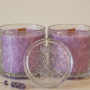 Peut inclure: Deux bougies en verre avec un motif floral, remplies de cire de couleur lavande. Les bougies ont des mèches en bois et un couvercle en verre transparent. Une brindille de lavande est au premier plan. Les bougies sont dans des récipients en verre transparent.