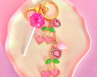 May include: A pink and gold cherry keychain with a lollipop charm. The keychain features a heart-shaped charm with rhinestones and a strawberry charm.