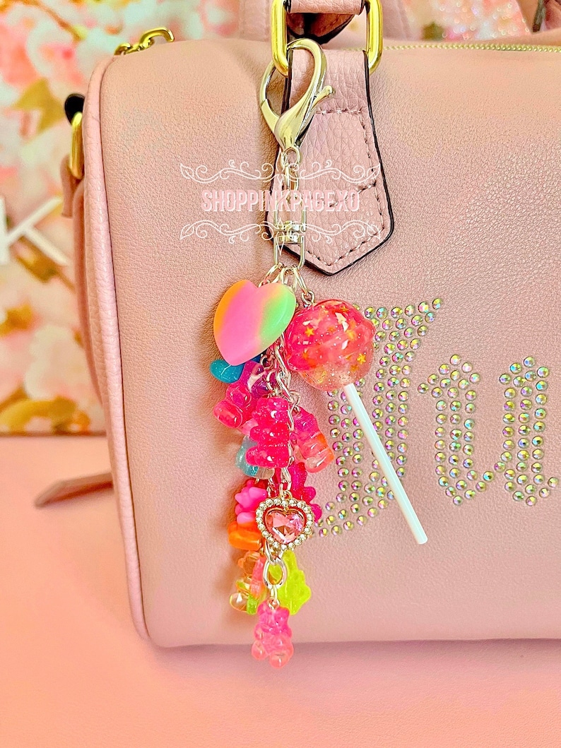 May include: A pink handbag with a decorative charm. The charm includes a rainbow heart, a pink lollipop, and colourful gummy bear charms. The handbag has the word "Juicy" spelled out in rhinestones.