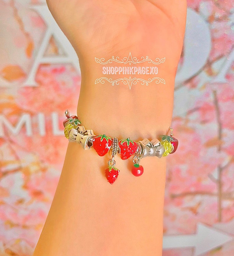 May include: A silver charm bracelet with red and green strawberry charms. The bracelet is on a person's wrist.