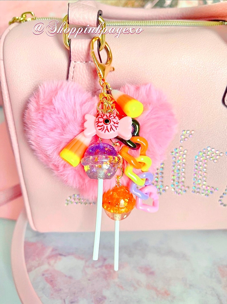K&ouml;nnte beinhalten: Ein rosa, flauschiger herzf&ouml;rmiger Anh&auml;nger mit einem goldenen Verschluss, verziert mit Zuckermais, einem Augapfel-Bonbon und zwei Lutscher-Anh&auml;ngern. Der Anh&auml;nger ist an einer rosa Handtasche mit dem Text "@Shoppinkpageco" befestigt.