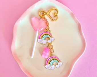 May include: A pink heart-shaped lollipop with a gold chain attached to a rainbow charm with white clouds and a pink gummy bear charm. The charm is on a white and gold dish with a pink background.