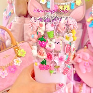 May include: A pink tumbler adorned with various charms, including bows, flowers, and food-themed trinkets. The tumbler has a pink straw and is surrounded by pink accessories. The text "Charming Cindy" is visible.