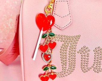 May include: A pink handbag with gold hardware and a charm featuring a red heart lollipop and cherries. The bag has the word "Juicy" written on it in gold rhinestones. The charm is attached to the bag's handle with a gold chain. The text "@Shoppinkpagexo" is visible on the bag.