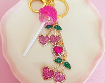 May include: A pink glitter lollipop charm with a gold key ring and a dangling charm of two pink glitter hearts with green leaves and a pink rhinestone heart.