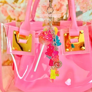 May include: A pink, transparent handbag with gold-coloured hardware and a charm. The charm features a lollipop, gummy bears, and a heart-shaped pendant. The text "SHOPPINGPAGEXO" is visible.