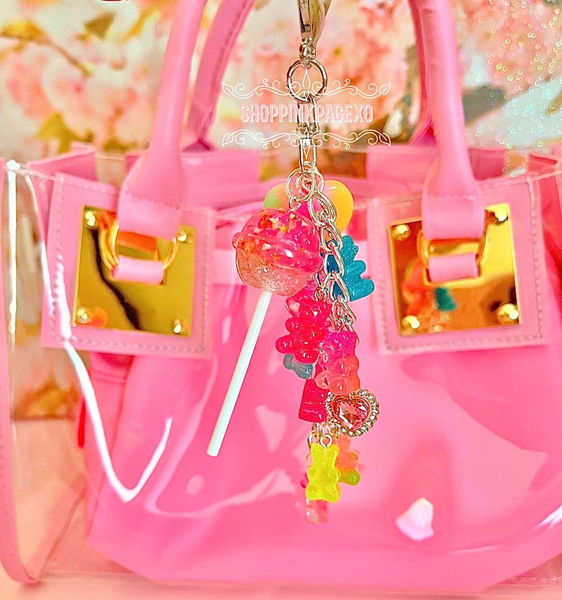 May include: A pink, transparent handbag with gold-coloured hardware and a charm. The charm features a lollipop, gummy bears, and a heart-shaped pendant. The text "SHOPPINGPAGEXO" is visible.