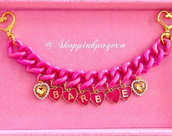 May include: Pink plastic chain charm bracelet with gold heart charms spelling out "BARBIE" and a gold heart clasp. The bracelet is in a pink velvet box.