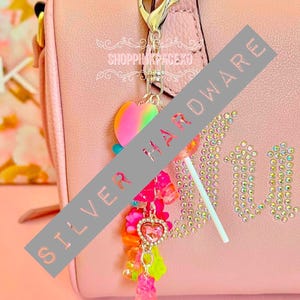 May include: A pink handbag with a silver clasp and a charm. The charm features a rainbow heart, a lollipop, and colourful beads and gummy bear charms. The word "Juicy" is spelled out in rhinestones.
