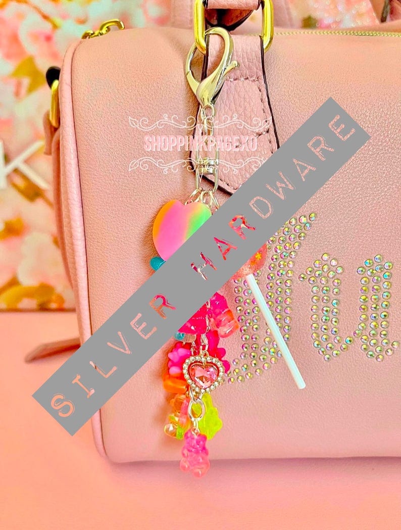May include: A pink handbag with a silver clasp and a charm. The charm features a rainbow heart, a lollipop, and colourful beads and gummy bear charms. The word "Juicy" is spelled out in rhinestones.