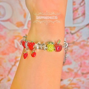 May include: A silver charm bracelet with a variety of fruit charms, including strawberries, apples, and pineapples. The charms are red, green, and yellow. The bracelet has a silver chain with a clasp.